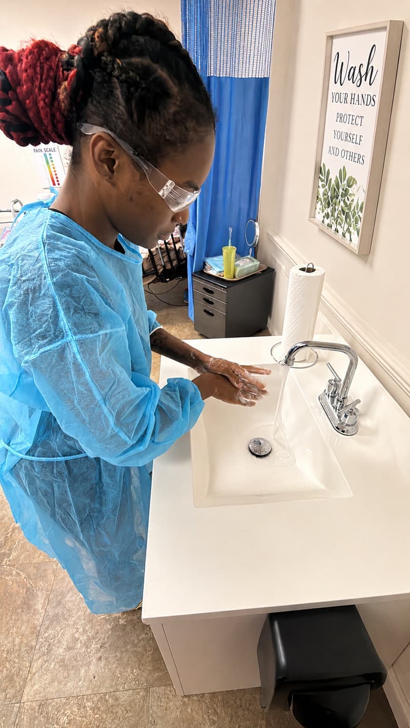 A CBRF student in a gown and safety glasses practising handwashing at the training sink.